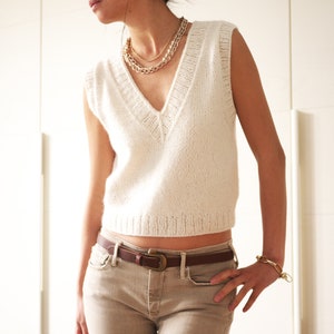 May include: A white sleeveless knitted sweater with a V-neckline. The sweater is worn over a brown belt with a gold buckle.