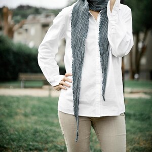 May include: A woman wearing a white button-down shirt with a long, blue and grey scarf tied around her neck. She is wearing beige trousers and standing in a grassy area.