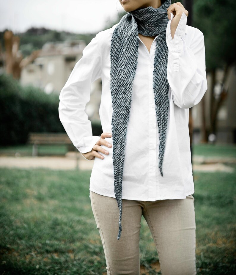 May include: A woman wearing a white button-down shirt with a long, blue and grey scarf tied around her neck. She is wearing beige trousers and standing in a grassy area.