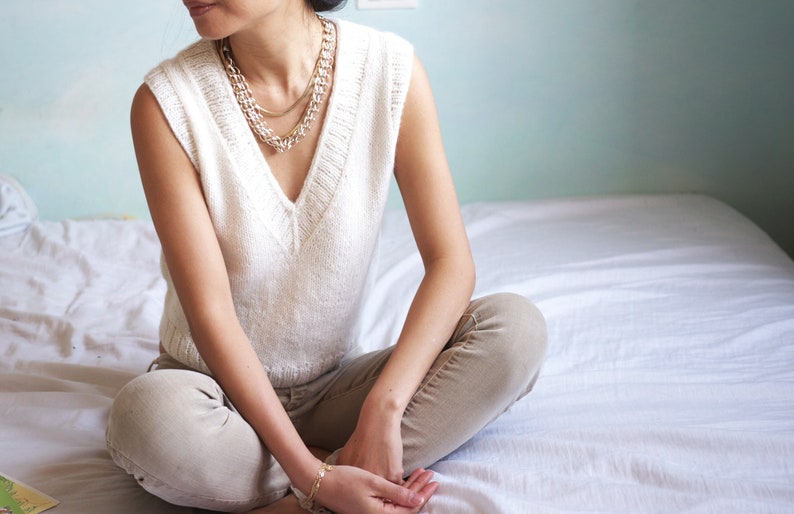 May include: A woman wearing a white sleeveless knitted sweater with a v-neck and multiple gold necklaces. She is sitting on a white bed with her legs crossed.