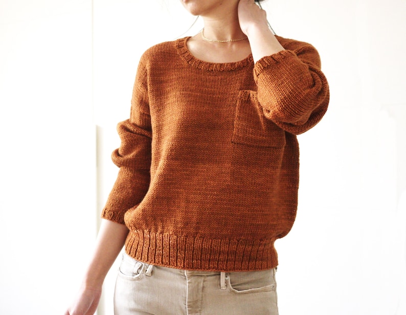May include: A brown knitted sweater with a patch pocket on the left side. The sweater has a round neckline and long sleeves.