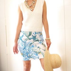 May include: A white sleeveless knitted top with a v-neckline and a white and blue floral skirt with a sea life design. The skirt features coral, starfish, and seaweed. The person is holding a straw hat.