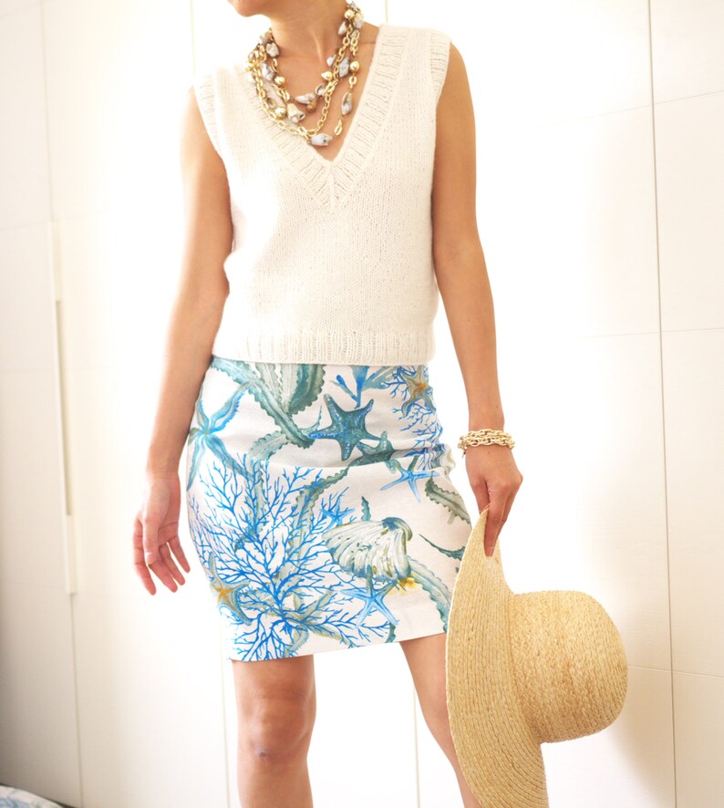 May include: A white sleeveless knitted top with a v-neckline and a white and blue floral skirt with a sea life design. The skirt features coral, starfish, and seaweed. The person is holding a straw hat.