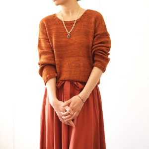 May include: A woman wearing a rust-colored knitted sweater with a gold chain necklace and a terracotta-colored skirt.