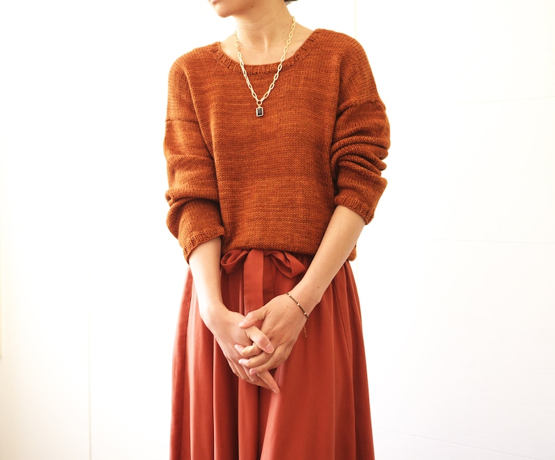 May include: A woman wearing a rust-colored knitted sweater with a gold chain necklace and a terracotta-colored skirt.