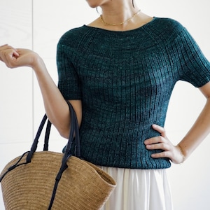 May include: A woman wearing a teal blue knitted top with a white skirt and a brown woven tote bag with black handles.