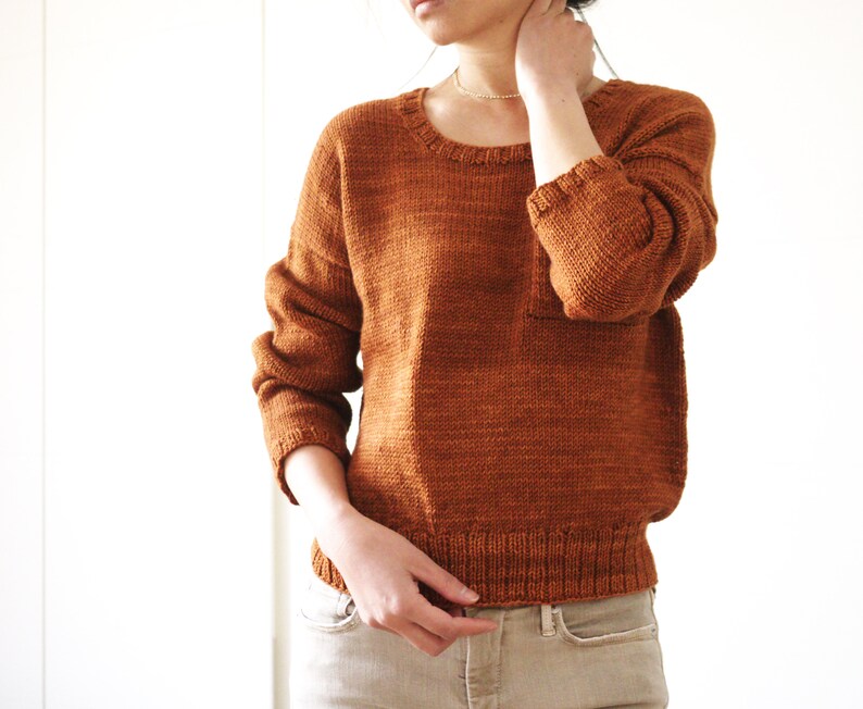 May include: A rust-colored knitted sweater with long sleeves and a crew neckline. The sweater is made of a soft, textured yarn and has a relaxed fit.