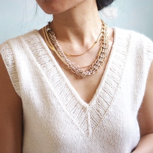 May include: A white sleeveless knitted sweater with a v-neck and a layered gold chain necklace.