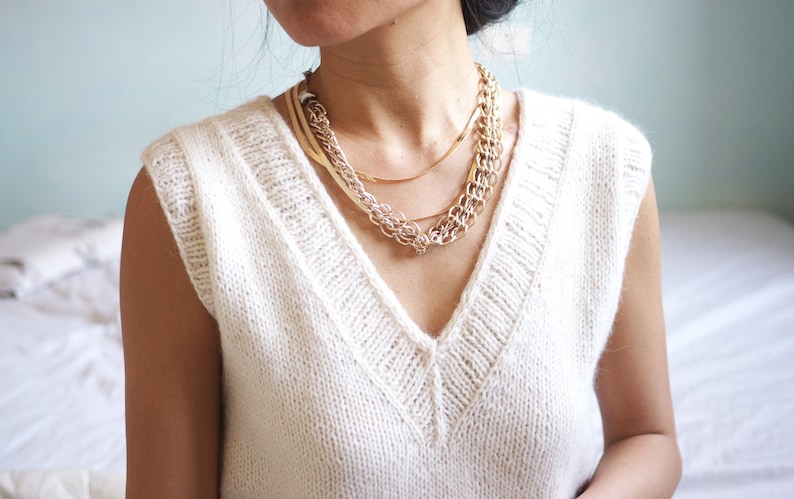 May include: A white sleeveless knitted sweater with a v-neck and a layered gold chain necklace.