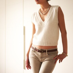 May include: A white knitted sleeveless sweater with a V-neckline and a brown leather belt. The sweater is cropped and the belt is fastened at the waist.