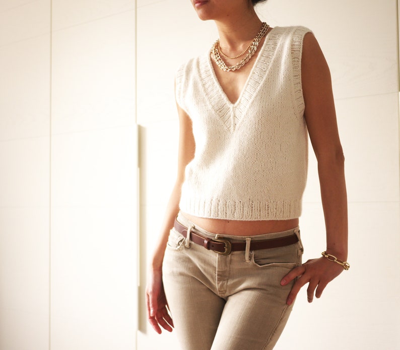 May include: A white knitted sleeveless sweater with a V-neckline and a brown leather belt. The sweater is cropped and the belt is fastened at the waist.