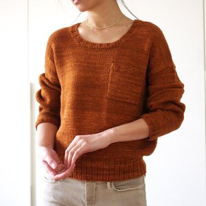 May include: A brown knitted sweater with a patch pocket on the front. The sweater has a round neckline and long sleeves.