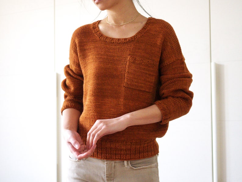 May include: A brown knitted sweater with a patch pocket on the front. The sweater has a round neckline and long sleeves.