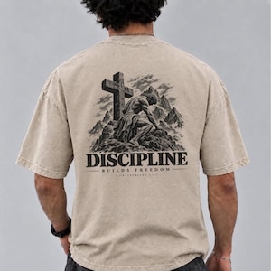 Könnte beinhalten: Ein beiges T-Shirt mit kurzen Ärmeln und einem schwarzen Aufdruck auf der Rückseite. Das Motiv zeigt eine Figur, die vor einem Kreuz in einer Berglandschaft kniet. Unter dem Bild steht der Text DISCIPLINE BUILDS FREEDOM und 1 CORINTHIANS 9:27.