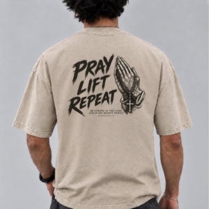 Op de afbeelding: Een beige t-shirt met korte mouwen en een zwarte afbeelding op de achterkant. Het ontwerp toont biddende handen die een rozenkrans met een kruis vasthouden, de tekst PRAY LIFT REPEAT en het vers BE STRONG IN THE LORD AND IN HIS MIGHTY POWER EPHESIANS 6:10.