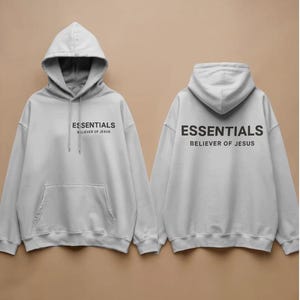 May include: Two light grey hoodies, each featuring the word "ESSENTIALS" in black print. The text "BELIEVER OF JESUS" is printed below. Both hoodies have a hood and a front pocket. The design is repeated on the front and back.