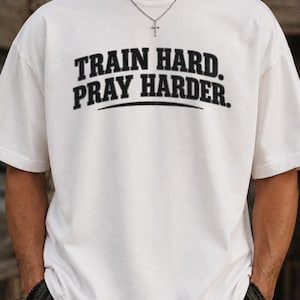 Op de afbeelding: Een wit T-shirt met korte mouwen en de zwarte tekst TRAIN HARD. PRAY HARDER. in een vetgedrukt, gebogen lettertype op de borst. Het shirt heeft een ronde hals en een relaxte pasvorm voor een strakke en eenvoudige uitstraling.