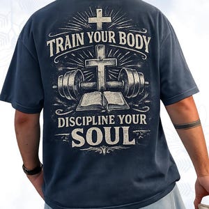 Könnte beinhalten: Ein dunkelblaues Kurzarm-T-Shirt mit einem Grafikdruck auf der Rückseite, der ein Kreuz, eine Langhantel und ein offenes Buch zeigt. Der Text "TRAIN YOUR BODY" steht über dem Kreuz und "DISCIPLINE YOUR SOUL" unter dem Buch in weißer Schrift.