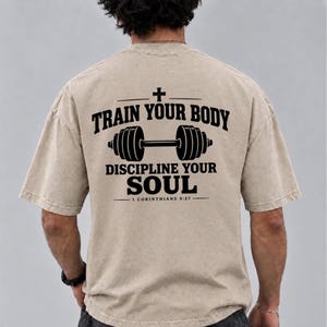 Könnte beinhalten: Ein beiges Kurzarm-T-Shirt mit schwarzem Text auf der Rückseite, der "TRAIN YOUR BODY DISCIPLINE YOUR SOUL 1 CORINTHIANS 9:27" lautet, mit einem Kreuzsymbol über dem Text und einer Hantelgrafik zwischen den beiden Hauptzeilen.