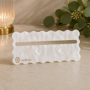 May include: A rectangular white acrylic stand with a wavy scalloped edge and a horizontal slot in the center. The surface has a pearlescent marbled texture. The text L'ATELIER RYAN is printed in gold on the bottom left corner of the stand.