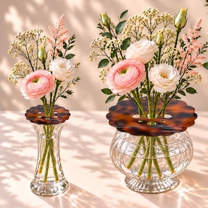 May include: Two clear glass vases with vertical ribbing and scalloped tortoise shell patterned lids. The vases contain arrangements of pink ranunculus, white flowers, and green foliage. The smaller vase is slender, while the larger is bulbous.