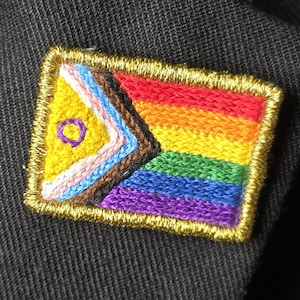 May include: Embroidered patch featuring a colorful flag design with a gold border. The flag incorporates rainbow stripes and other geometric shapes. The patch is attached to a black fabric, likely a jacket or blazer.