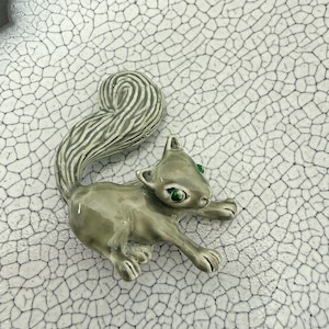 May include: A gray squirrel figurine with green eyes. The squirrel is in a crouching position, with its tail curled upwards. The figurine is set against a white, cracked ceramic surface.