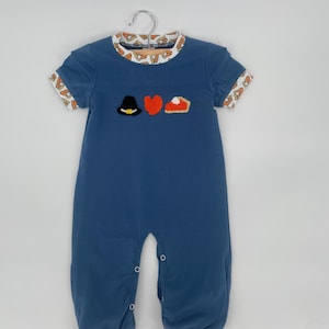 May include: A blue baby romper with short sleeves and a round collar. The collar and sleeve cuffs have a patterned trim. The romper features embroidered images of a pilgrim hat, a heart, and a slice of pie. The romper is hanging on a metal hanger.