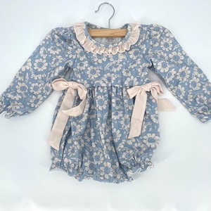 May include: A baby romper with a blue floral pattern and long sleeves. The romper features a ruffled collar and two pink bows at the waist. The garment is hanging on a wooden hanger.