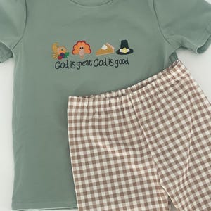 May include: Sage green short-sleeved t-shirt with embroidered Thanksgiving-themed graphics and the text "God is great God is good." Paired with brown and white checkered leggings. The t-shirt has a small tag that says "Little Charlee Lane."
