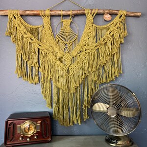 May include: A large, olive-green macrame wall hanging suspended from a wooden branch. The intricate design features knotted patterns and fringe. A vintage-style radio and a metal fan are in the foreground, creating a retro aesthetic.