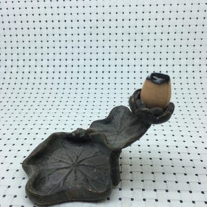 Pode incluir: Um porta-incenso de cor bronze em forma de folha de lótus. O suporte tem um pequeno cone de incenso castanho com a parte superior preta. O design inclui uma bandeja em forma de folha e uma folha curva que suporta o cone.