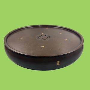 Puede incluir: Un recipiente redondo de color marrón oscuro con tapa y acabado mate liso. La tapa presenta un emblema decorativo central y motas de color dorado dispersas por la superficie. El recipiente tiene una base baja en forma de cuenco con tapa a juego.
