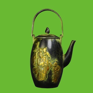 May include: A dark metal teapot with a swing handle and a lid featuring a finial. The body is decorated with a raised gold-toned relief depicting a seated figure holding a staff, accompanied by two smaller figures, all set against a dark patina.