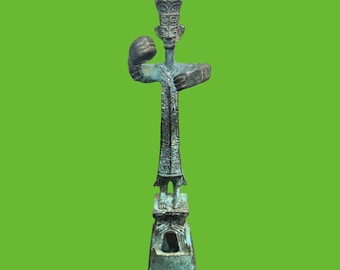 May include: A bronze-colored, stylized sculpture with a figure holding a fist and a rectangular object. The figure has a detailed face and is atop a long, decorated column. The sculpture has a green patina and sits on a rectangular base.