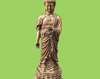 May include: A bronze-colored standing Buddha statue featuring a textured ushnisha, draped robes with flowing folds, and a manji symbol on the chest. The figure stands on a circular lotus-shaped pedestal with detailed petals, captured in a full-length view.