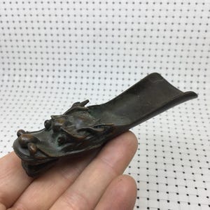 May include: A bronze-colored, dragon-shaped incense burner. The dragon's head is detailed with scales and features, and the burner has a long, curved trough. The object is held in a hand against a dotted background.