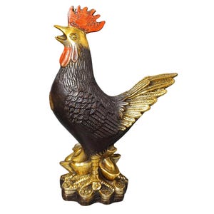 May include: A bronze-colored rooster statue with gold accents. The rooster has a red comb and wattle, with detailed feathering. It stands on a base of gold coins and ingots, with its beak open as if crowing.