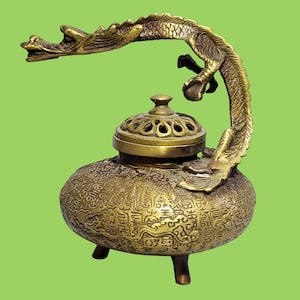 May include: A brass incense burner featuring a rounded body covered in intricate embossed characters and patterns. The vessel stands on three short legs and includes a removable lid with cut-out details, topped by a curved dragon handle.