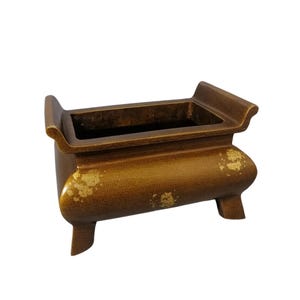 May include: A bronze-colored incense burner with a rectangular basin and curved sides. The burner has three short legs and decorative gold leaf accents. The top edges are slightly raised, and the overall design is traditional.
