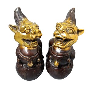Può includere: Due statue color bronzo di creature mitologiche con teste color oro. Le creature hanno la bocca aperta, denti aguzzi e capelli a spillo. Indossano abiti ornati con accenti dorati. Le statue sono su sfondo bianco.