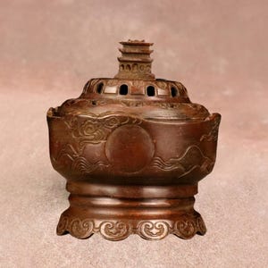 May include: A bronze incense burner with detailed carvings. The round body features a wave and circle pattern. The lid has several holes and a pagoda-style top. The base has an ornate design. The overall design is intricate and decorative.