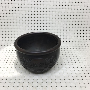 May include: A dark brown, round bowl with decorative carvings around the outside. The bowl has a wide opening and a smooth, dark interior. The carvings feature a traditional Asian design. The bowl is on a white woven surface.