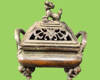 May include: A dark bronze-toned metal incense burner with a square base, two curved side handles, and four legs shaped like figures. The lid features an openwork floral pattern and a sculpted mythical creature standing on a ball at the top.