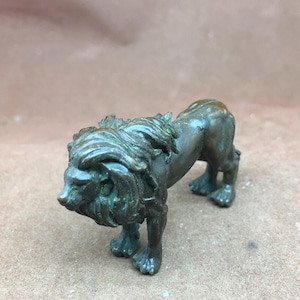 Könnte beinhalten: Kleine Löwenfigur aus Bronze. Der Löwe hat eine detaillierte Mähne und einen muskulösen Körper, in einer Laufhaltung mit gesenktem Kopf. Die Bronze hat ein verwittertes Aussehen mit grünen Patina-Akzenten.