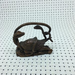 Pode incluir: Uma escultura de cor bronze com uma tartaruga e duas cobras. Uma cobra está enrolada na tartaruga, e a outra arqueia-se sobre ela. A escultura está sobre uma base texturizada.