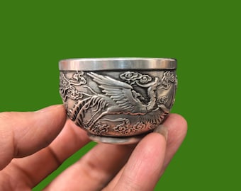 May include: A small, silver-colored cup with an engraved phoenix design. The cup is held in a hand against a green background. The phoenix is depicted with outstretched wings amidst clouds, showcasing detailed craftsmanship.