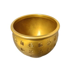 Op de afbeelding: Een goudkleurige, ronde kom met een brede rand. De buitenkant van de kom is versierd met in reliëf aangebrachte Chinese karakters. De kom heeft een glad, reflecterend oppervlak en staat tegen een effen witte achtergrond.