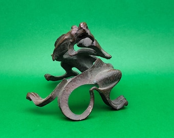 May include: A bronze sculpture of a stylized fish leaping over a wave. The sculpture has a dark, aged patina and is set against a green background. The fish's form is detailed, with visible fins and scales.
