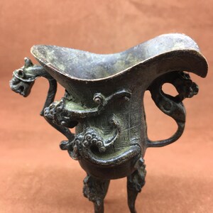 Puede incluir: Antiguo recipiente de bronce con borde acampanado y tres patas. El recipiente presenta intrincados diseños de dragones y volutas en las asas y el cuerpo. El bronce tiene una pátina envejecida, dándole un aspecto vintage.
